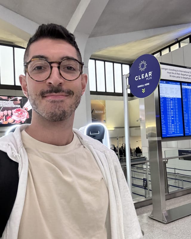 Rocky standing beside a round CLEAR Plus enroll-here sign at an airport checkpoint, departure boards glowing behind him