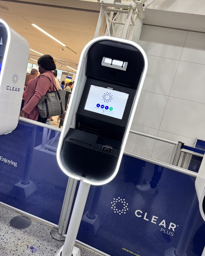 A CLEAR pod seen from the side, its lit white ring framing a dark screen above the blue CLEAR Plus barrier, November 2023