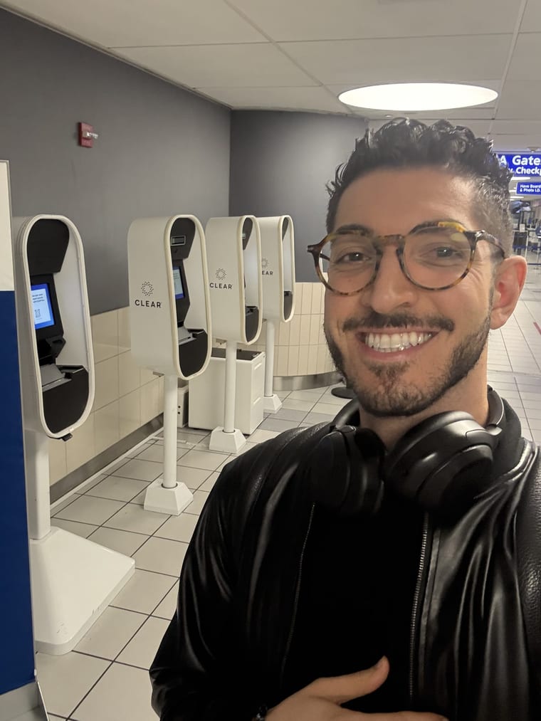 Rocky smiling in front of a row of older stop-and-scan CLEAR pods in a quiet checkpoint area, May 2025